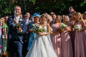 My signature photograph. A groom celebrates his wedding by spraying a bottle of champagne beside his bride and in front of his family and friends. Devon Wedding Photographer. GoodLuckPhotography