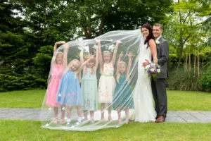 Newlywed couple stand holding hands whilst five young girls play under her veil with their arms in the air, lots of smiles and laughter by Devon photographer - robb webb