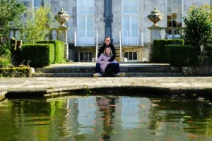 North Devon Photographer - Robb Webb A couple pose for pictures beside a large pond full of orange carp fish beside a country house during their pre wedding meeting with Devon wedding Photographer Robb Webb of good luck photography. A picture of a brides Shoes, lipstick and jewellery on a wooden table top in the bridal suite of a country house hotel with sunlight streaming across the image from the right hand side. Devon Wedding Photographer. A picture of the flying lady mascot on the Rolls Royce limousine car that chauffered the bride to her wedding day. The mascot has the wedding ribbons wrapped around its base and there are bokeh in the shot. Devon wedding Photographer. GoodLuckPhotography